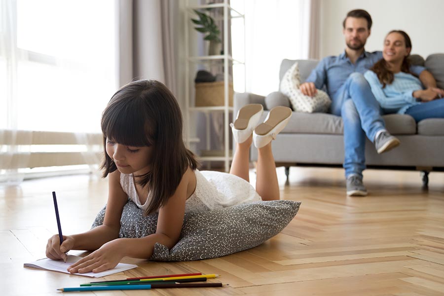 About Our Agency - Family in the Living Room at Home with Young Daughter Lying on a Soft Cushion on the Floor with Parents Sitting on the Sofa