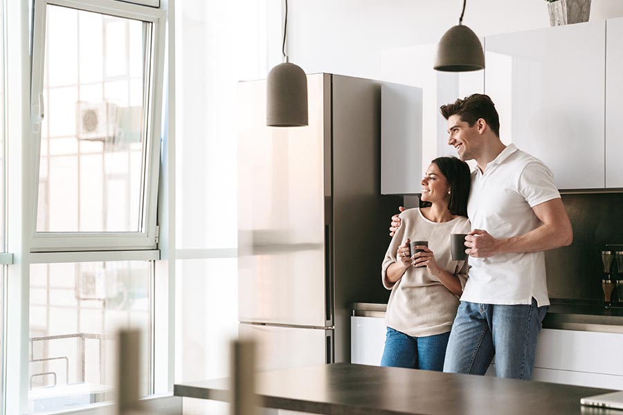 Personal Insurance - Happy Young Couple Holding Coffee Mugs and Looking Out the Window of a Modern Kitchen in Their New Home